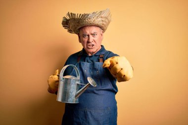 Orta yaşlı çiftçi önlük ve şapka takıyor sarı arka plan üzerinde sulama tenekesi sinirlenmiş ve sinirlenmiş öfkeyle bağırıyor ve el kaldırarak bağırıyor. Öfke kavramı.