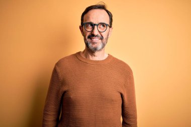 Orta yaşlı, beyaz, kahverengi süveter ve gözlük takan, izole edilmiş sarı arka planda gülümseyen ve kafasını çeviren bir adam..