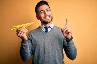 Elinde kağıt uçak tutan genç yakışıklı adam izole edilmiş sarı arka planda bir fikir ya da soru işareti ile şaşırdı.