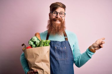 Kızıl saçlı İrlandalı süpermarket çalışanı, sakallı, önlüklü ve elinde taze yiyeceklerle elini ve parmağını kenarı göstererek çok mutlu.