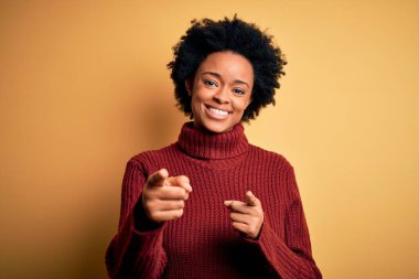 Kıvırcık saçlı, balıkçı yaka kazaklı, neşeli ve komik yüzlü genç Afro-Amerikan kadın kamerayı işaret ediyor. İyi enerji ve titreşimler.