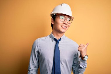 Genç, yakışıklı, Çinli mimar, güvenlik kaskı takmış ve sarı arka planda kravat takmış. Gülümsüyor, gülümsüyor ve başparmağıyla yan tarafı işaret ediyor..