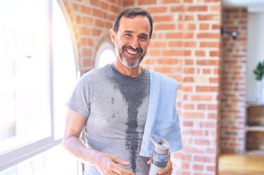 Orta yaşlı yakışıklı sporcu, spor salonunda spor salonunda bir şişe su ve havlu tutarak terliyor. Mutlu bir yüzle ayakta duruyor ve gülümseyerek dişlerini gösteriyor.