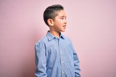 Zarif bir gömlek giyen küçük bir çocuk pembe izole edilmiş arka planda durup yüzünde gülümsemeyle, doğal bir ifadeyle yan yana bakıyor. Kendinden emin gülen.