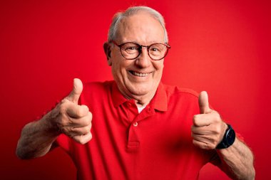 Gri saçlı, gözlüklü ve kırmızı arka plan tabelasının üzerine gündelik tişört giyen, el hareketleriyle olumlu hareket eden, gülümseyen ve mutlu bir adam. Neşeli ifade ve kazanan jest.