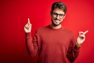 Sakallı, gözlüklü ve süveterli, kırmızı arka planda duran, kendine güvenen, farklı yönleri gösteren genç, yakışıklı bir adam. Reklam için boşluk kopyala