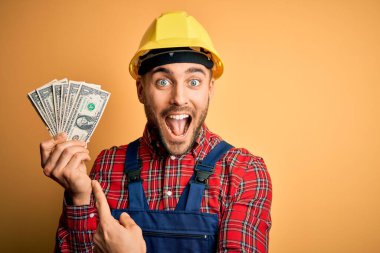 Güvenlik kaskı takan genç müteahhit, sarı arka planda dolar karşılığı para tutuyor. Elle ve parmakla göstererek çok mutlu.