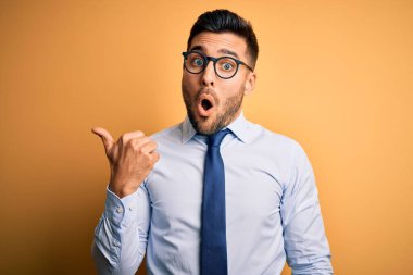 Sarı arka planda duran kravat ve gözlük takan genç yakışıklı işadamı el parmağı yana dönük, ağzı açık şaşkın bir ifadeyle şaşırdı..