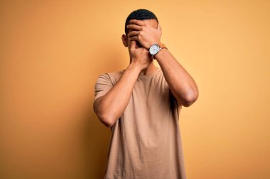 Genç, yakışıklı, Afro-Amerikalı bir adam sarı arka plan üzerinde dikilmiş gözleri ve ağzı kapalı, şaşırmış ve şok olmuş bir şekilde duruyor. Duygularını gizliyor.