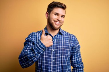 Sarışın, sakallı ve mavi gözlü, sarı arka planda gömlek giyen genç bir işadamı mutlu bir el hareketi yapıyor. Kameraya bakıp başarıyı gösteren ifade onaylanıyor.