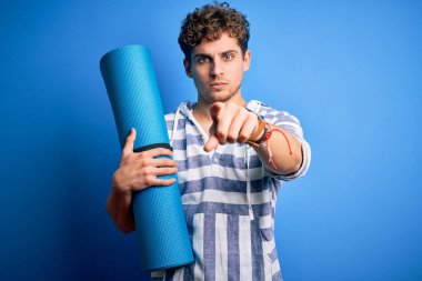 Sarışın, kıvırcık saçlı, yoga yapmak için minder tutan genç bir adam izole edilmiş mavi arka planda parmağını kameraya doğrultuyor ve sen, el işareti, ön taraftan pozitif ve kendinden emin bir hareket.
