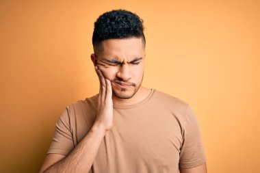 İzole edilmiş sarı arka planda dikilen sıradan tişört giyen genç yakışıklı diş ağrısı ya da diş ağrısı yüzünden ağız ve acı dolu yüz ifadelerine dokunuyor. Dişçi