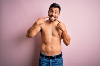 Sakalsız, genç, yakışıklı, güçlü bir adam pembe arka planda dikiliyor. Gülümsüyor, parmaklarıyla ve ağzıyla işaret ediyor. Diş sağlığı konsepti.