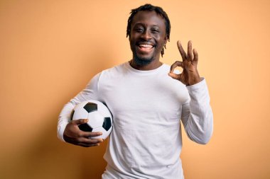 Genç Afrikalı Amerikalı oyuncu futbol topunu sarı arka planda tutarak oynuyor. Tamam işareti yapıyor, mükemmel bir sembol.