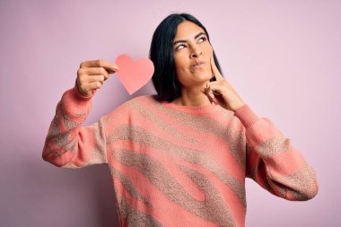 Genç, güzel, İspanyol bir kadın pembe arka planda romantik bir kalp kâğıdı tutuyor. Ciddi bir yüz. Soruyu düşünüyor, kafası karışık bir fikir.
