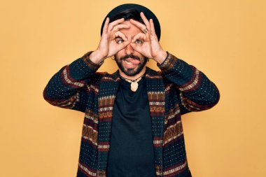 Genç, yakışıklı, bohem, hippi tarzı giyinmiş ve bohem şapkalı bir adam dürbün gibi jest yapıyor, gözleri parmaklarından bakıyor. Çılgın ifade.