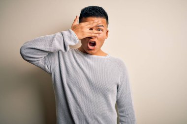 Genç, yakışıklı, sıradan bir süveter giyen, izole edilmiş beyaz arka planda dikilen, şok içinde, yüzünü ve gözlerini kapayarak utanmış bir ifadeyle parmaklarının arasından bakan Latin bir adam..