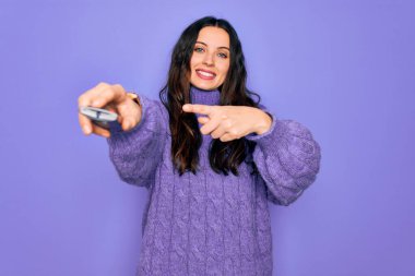 İzole edilmiş mor arka plan üzerinde uzaktan kumanda kullanan güzel bir kadın. Elle ve parmakla işaret ederek çok mutlu.