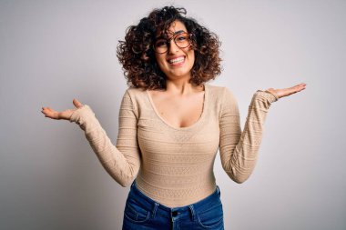 Genç, kıvırcık, kıvırcık Arap bir kadın beyaz arka planda günlük tişört ve gözlük takıyor. Gülümsüyor, avuçlarını açıyor, sunuyor, reklam yapıyor, karşılaştırıyor ve dengede duruyor.