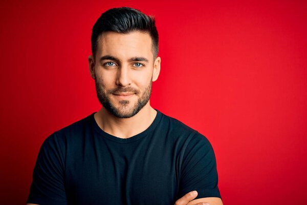 Young handsome man wearing casual black t-shirt standing over isolated red background happy face smiling with crossed arms looking at the camera. Positive person.