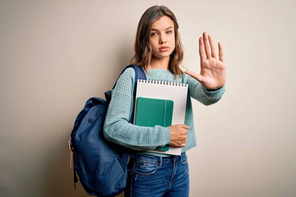 Young blonde student girl wearing backpack and books from school over isolated background with open hand doing stop sign with serious and confident expression, defense gesture