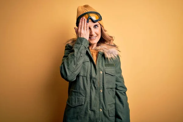 中年美丽的金发滑雪者女人 身穿雪地运动鞋 戴着雪地护目镜 一只手捂住眼睛 脸上挂着自信的微笑 还有令人惊讶的情绪 — 图库照片