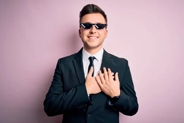 Young handsome caucasian business man wearing funny thug life glasses smiling with hands on chest with closed eyes and grateful gesture on face. Health concept.