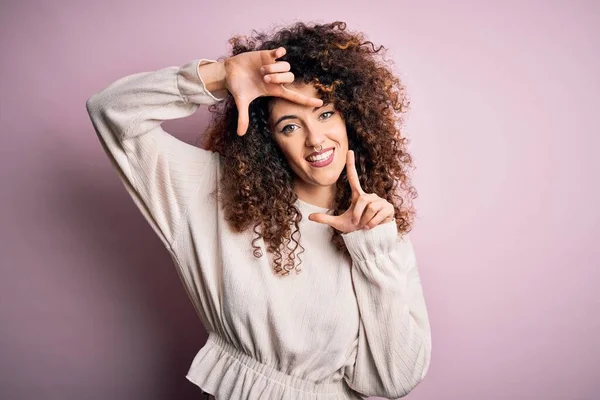 カールした髪とピアスをした美しい女性ピンクの背景にカジュアルなセーターを身に着けている笑顔幸せな顔を持つ手や指でフレームを作ります 創造性と写真の概念 — ストック写真