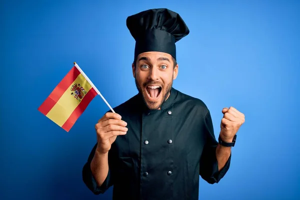 Young handsome cooker man with beard wearing uniform holding spain spanish flag screaming proud and celebrating victory and success very excited, cheering emotion