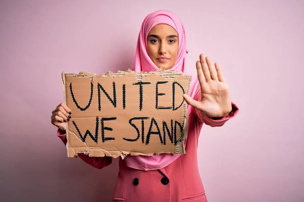 Young activist woman wearing pink muslim hijab holding banner with united we stand message with open hand doing stop sign with serious and confident expression, defense gesture