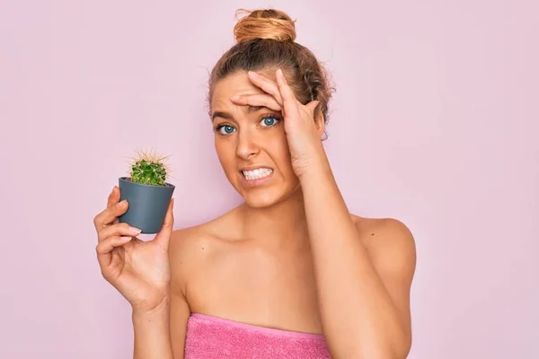 Beautiful blonde woman with blue eyes wearing towel shower after bath holding small cactus stressed with hand on head, shocked with shame and surprise face, angry and frustrated. Fear and upset for mistake.