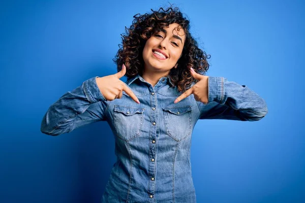 年轻美丽的卷曲阿拉伯女人 穿着休闲装 站在蓝色的背景上 面带微笑 用手指指着自己 自豪而快乐 — 图库照片