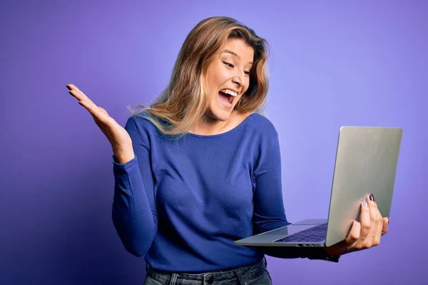 年轻美丽的金发女子在紫色的背景下 用笔记本电脑工作 她非常快乐而兴奋 胜利的表情带着大大的微笑和举手庆祝胜利的胜利 — 图库照片