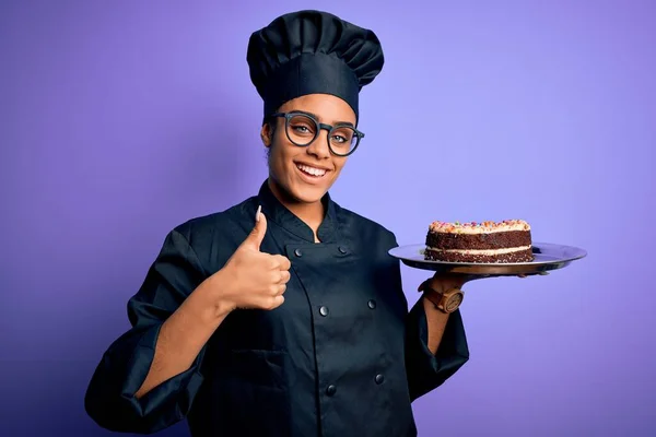 Young african american cooker girl wearing uniform and hat holding tray with cake happy with big smile doing ok sign, thumb up with fingers, excellent sign