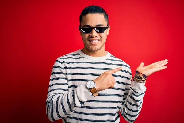 Young brazilian man wearing funny thug life sunglasses over isolated red background amazed and smiling to the camera while presenting with hand and pointing with finger.