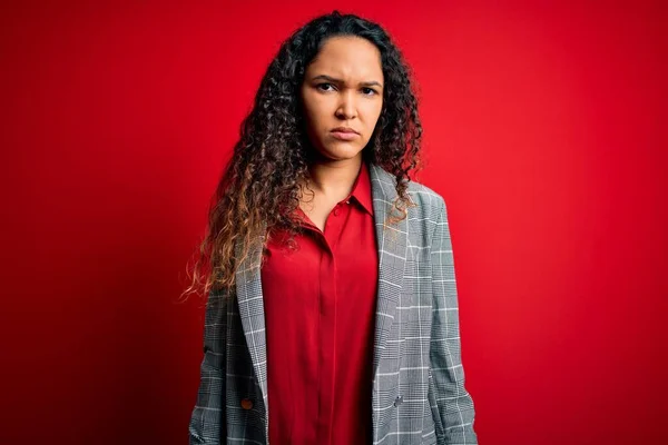 Young beautiful businesswoman with curly hair wearing elegant jacket over red background with serious expression on face. Simple and natural looking at the camera.