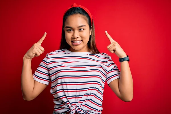 stock image Young beautiful asian girl wearing casual striped t-shirt over isolated red background smiling pointing to head with both hands finger, great idea or thought, good memory