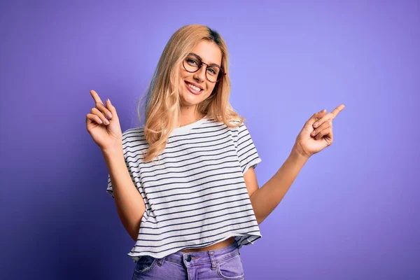 年轻美丽的金发女人穿着条纹T恤 戴着紫色背景的眼镜 带着自信的微笑 手指指向不同的方向 广告的复制空间 — 图库照片