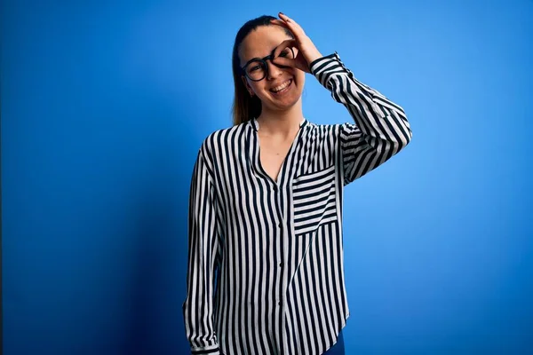 Beautiful blonde woman with blue eyes wearing striped shirt and glasses over blue background doing ok gesture with hand smiling, eye looking through fingers with happy face.