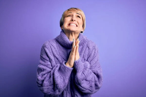 Young blonde woman with short hair wearing winter turtleneck sweater over purple background begging and praying with hands together with hope expression on face very emotional and worried. Begging.
