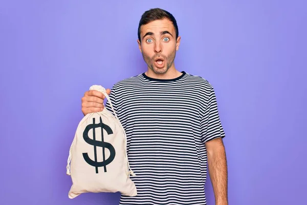 Young handsome man with blue eyes holding bag with dollar symbol over purple background scared in shock with a surprise face, afraid and excited with fear expression