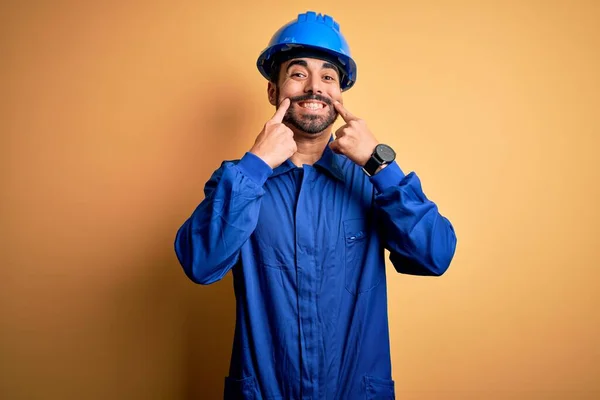Mechanic man with beard wearing blue uniform and safety helmet over yellow background Smiling with open mouth, fingers pointing and forcing cheerful smile