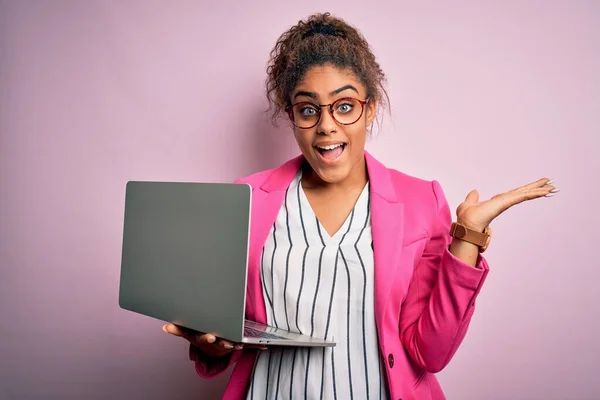 African american business woman wearing glasses working using laptop over pink background very happy and excited, winner expression celebrating victory screaming with big smile and raised hands