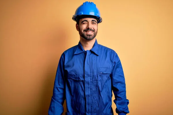 機械的な男で髭を身に着けています青い制服と安全ヘルメット上の黄色の背景ウィンク見ますカメラでセクシー式 陽気で幸せな顔 — ストック写真