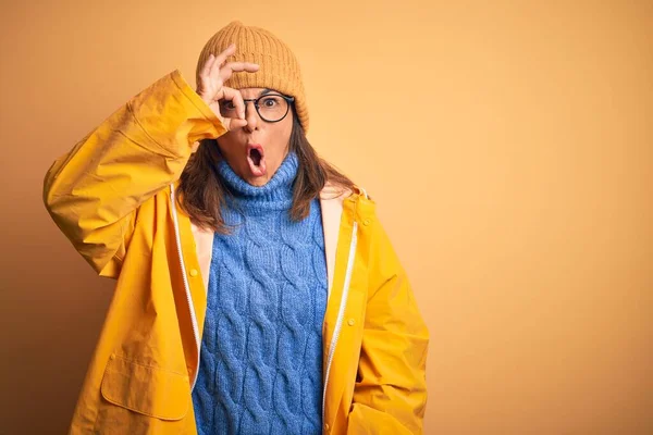 Middle age woman wearing yellow raincoat and winter hat over isolated background doing ok gesture shocked with surprised face, eye looking through fingers. Unbelieving expression.