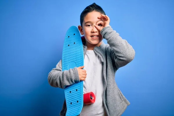 小男孩滑板手抱着现代滑板在蓝色孤立的背景上 笑容满面 手牵手看着手指 做好手势 — 图库照片