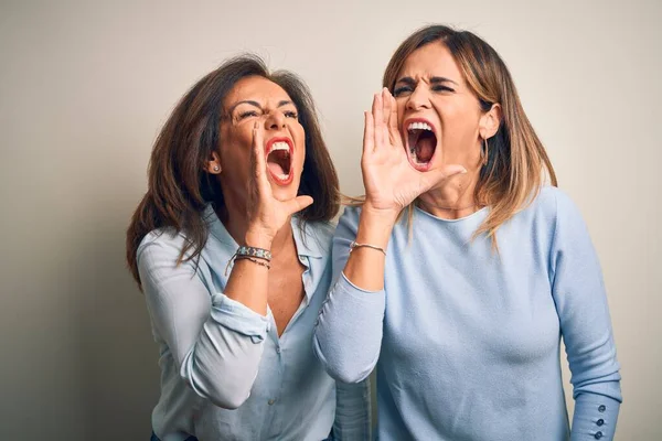 Middle age beautiful couple of sisters standing over isolated white background shouting and screaming loud to side with hand on mouth. Communication concept.
