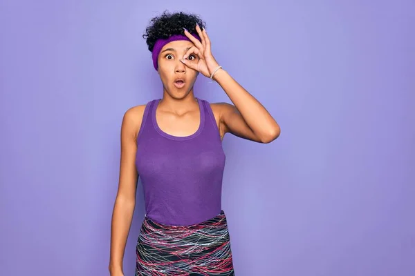 Beautiful african american afro woman wearing casual sportswear over purple background doing ok gesture shocked with surprised face, eye looking through fingers. Unbelieving expression.