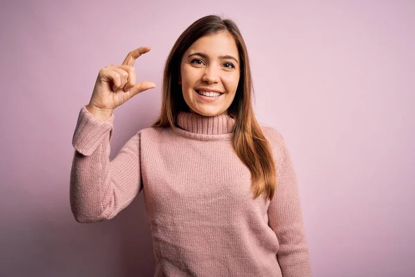 ピンクの隔絶された背景にタートルネックセーターを身に着けている美しい若い女性は笑顔と指を見て カメラと小さなサイズの看板を行う手で自信を持ってジェスチャー 概念の測定 — ストック写真