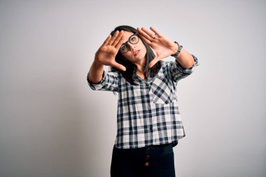 Mavi gözlü, beyaz arka planda günlük gömlek ve gözlük takan, el ve parmaklarını kullanarak çerçeve yapan esmer bir kadın.